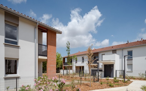 Résidence Le Floréal de logements neufs composée de maisons mitoyennes blanches à toiture en tuiles rouges, avec allées piétonnes, jardins paysagers et ciel bleu partiellement nuageux.