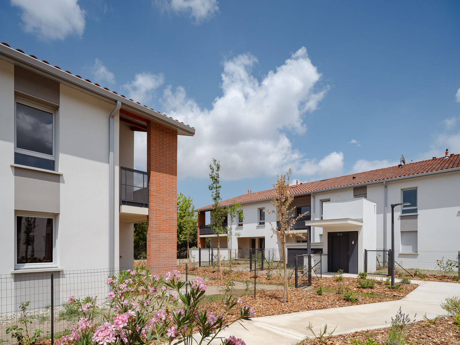 Résidence Le Floréal de logements neufs composée de maisons mitoyennes blanches à toiture en tuiles rouges, avec allées piétonnes, jardins paysagers et ciel bleu partiellement nuageux.