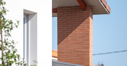 Détail d’un bâtiment résidentiel contemporain avec façade blanche, pilier en brique rouge, balcon à garde-corps métallique gris et toiture en tuiles, entouré de végétation sous un ciel bleu dégagé.