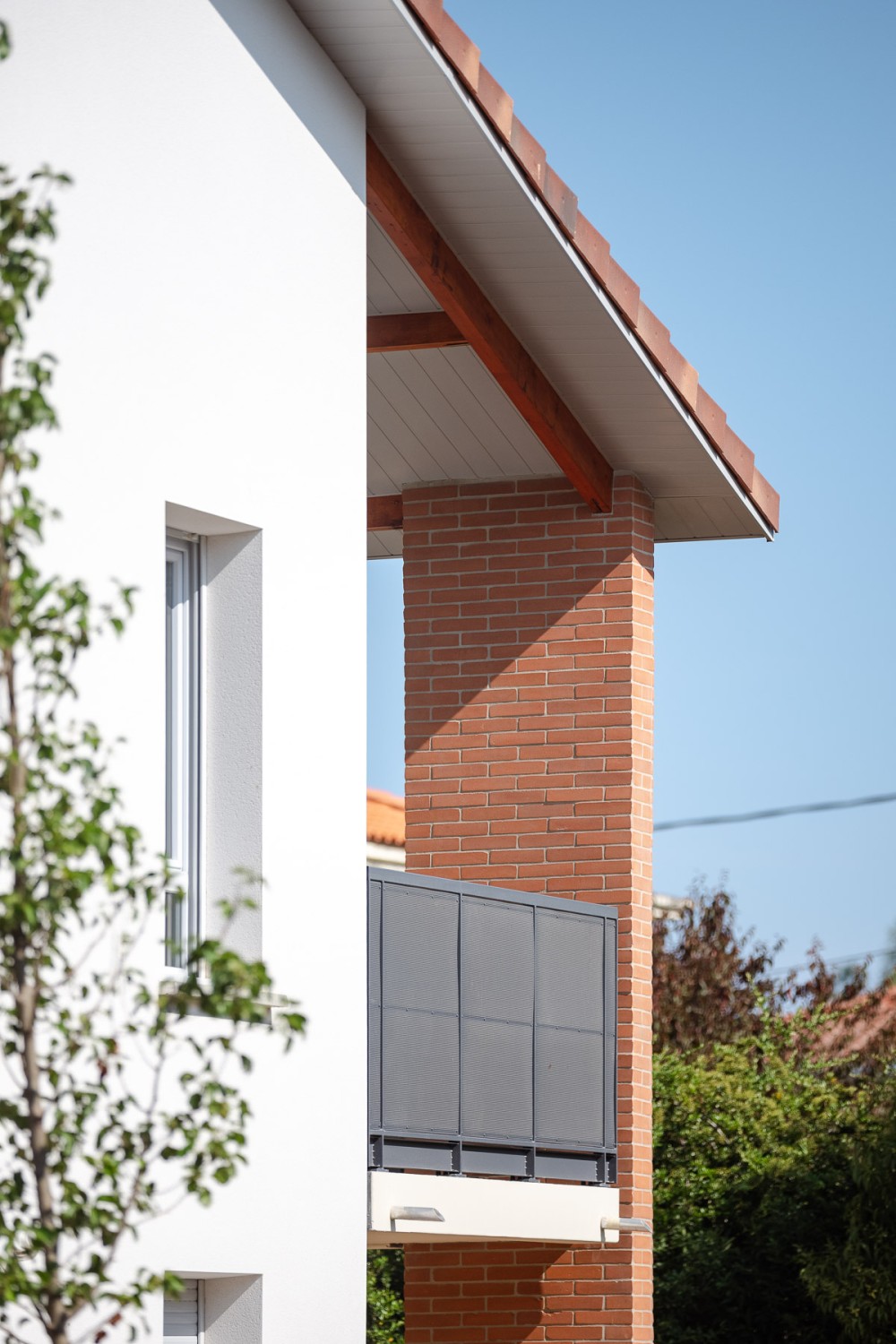 Détail d’un bâtiment résidentiel contemporain avec façade blanche, pilier en brique rouge, balcon à garde-corps métallique gris et toiture en tuiles, entouré de végétation sous un ciel bleu dégagé.
